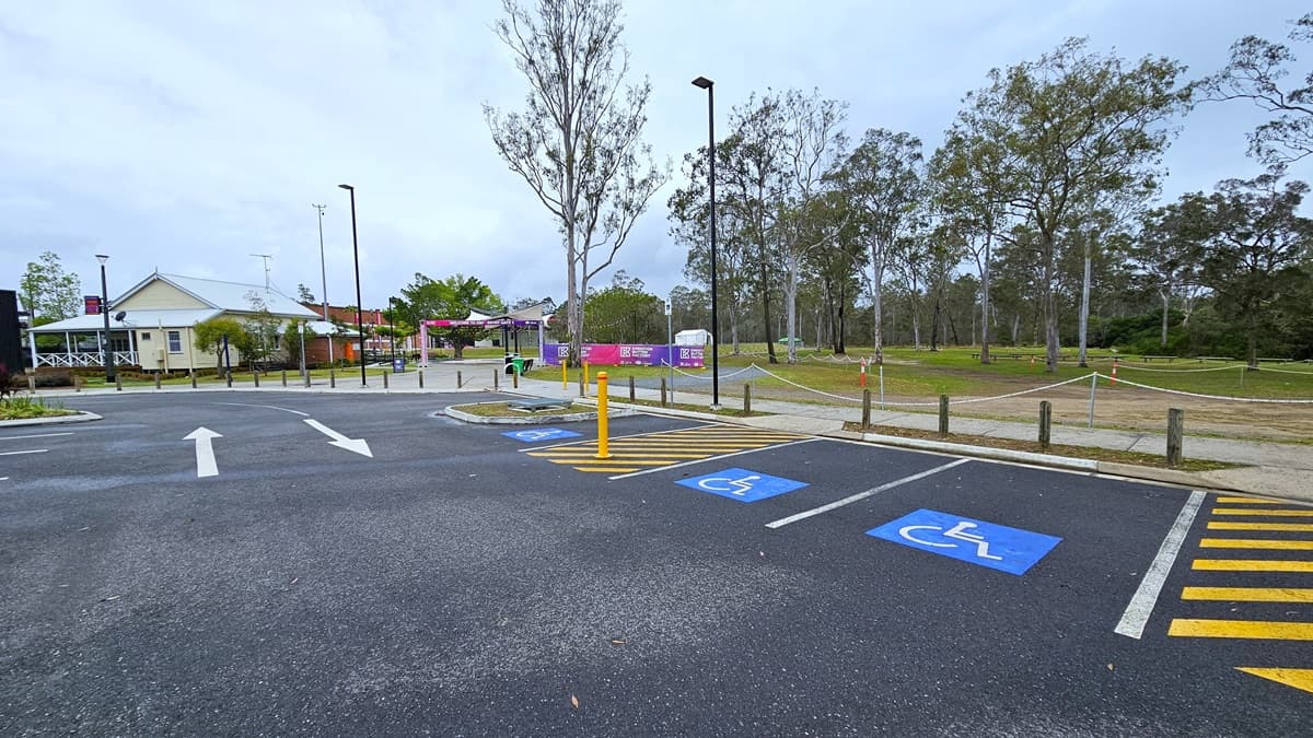 The parking area includes three accessible spaces on the right, marked with blue wheelchairs and yellow diagonal lines. A low rope fence, concrete bollards, and tall lamp posts line the edge, while a concrete plant box is positioned on the right. Trees and grass surround the area, leading to a white and brick building with colorful banners displaying a gradient shade of pink and blue.