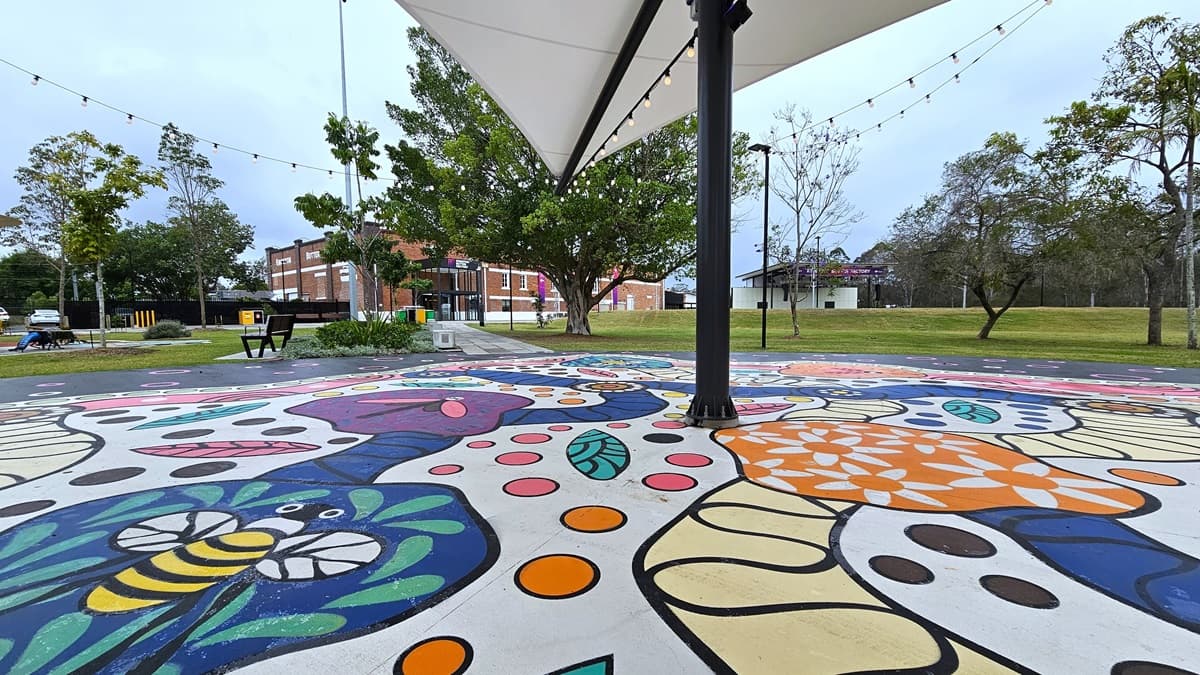 In the center is a shade structure with light bulbs. Underneath, a colorful floor painting depicts imagery of bees, dragonflies, flowers, and leaves. On the left, a bench is positioned near a landscape plant box. Further ahead, a concrete pathway leads to a brick-red building. Tall trees and grass surround the area.