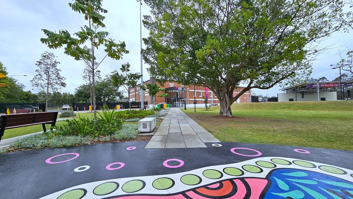 In the middle is a floor with colorful prints, connected to a concrete path leading to the brick-red building. On the left, a sitting bench is surrounded by ornamental plants and trees. Stone structures with inscriptions are lined up on the left. To the right, a tall tree with robust branches stands, and further right is a stage. Green grass surrounds the area on both sides.