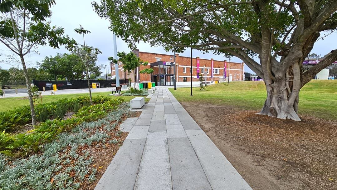In the middle is a concrete path leading to the red brick building adorned with gradient banners. On the left, there's a landscape of ornamental plants and trees, with a bench for sitting further ahead. Stone structures with inscriptions are lined up on the left. To the right, a tall tree with robust branches stands, and further right is a stage. Green grass surrounds the area on both sides.