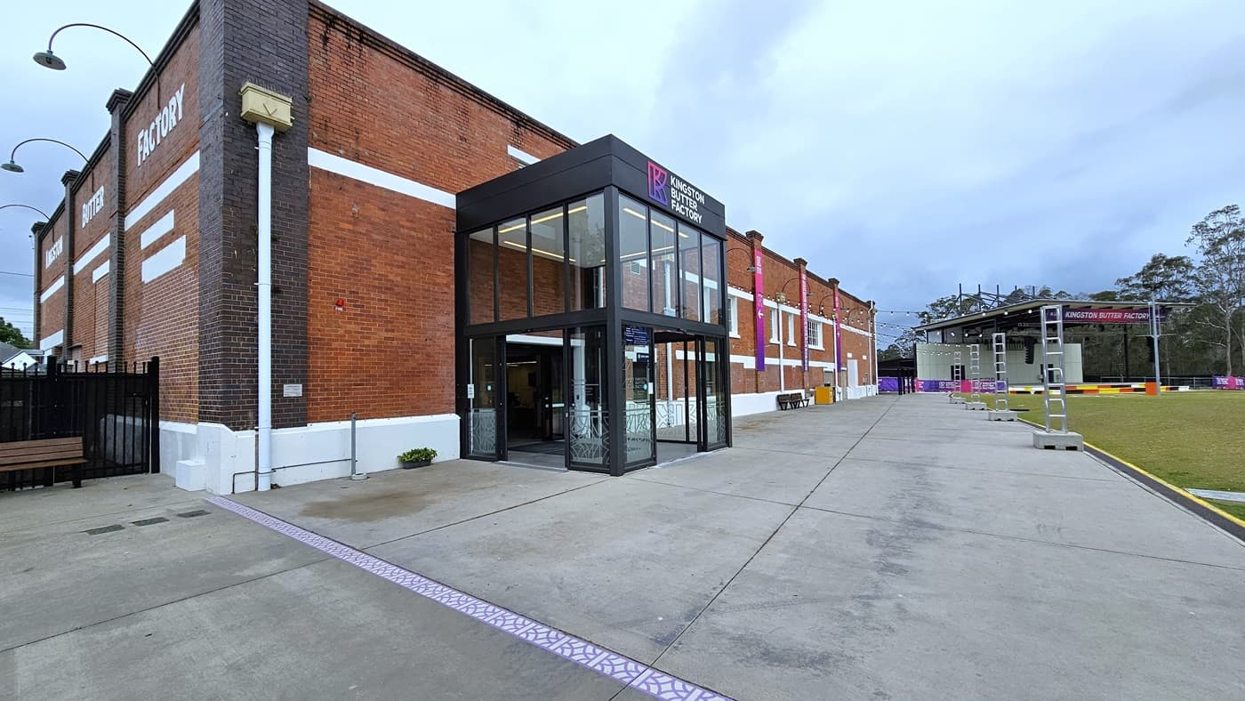 In the middle is an industrial building constructed from red bricks, featuring glass windows. The front is decorated with a gradient blue and pink banner, vertically suspended on the right alongside light fixtures. The entrance, a double modern glass and steel structure, displays facility signage above the glass door. On the left, there's a black steel fence with a bench for seating. To the right, a stage is adorned with gradient banners, and nearby, there's a grassy area with steel structures.