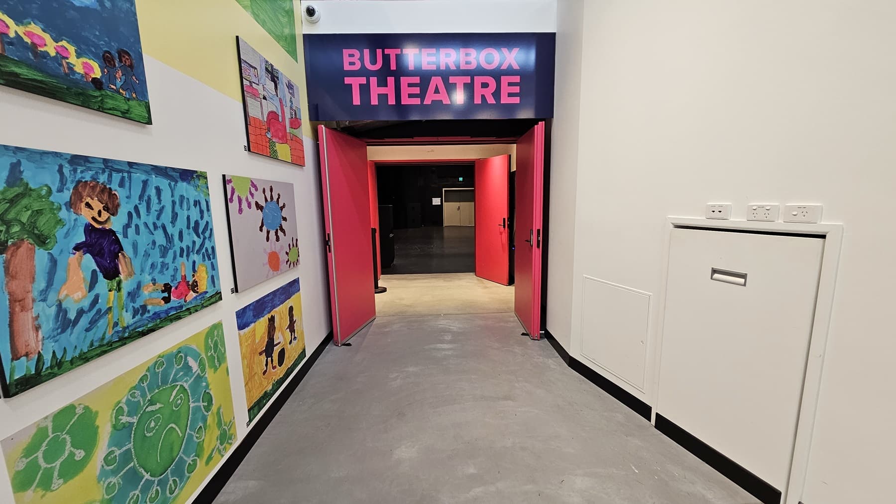 To the left, a wall features adorned art paintings. In the middle, a bright pink door opens outward, accompanied by an overhead sign displaying "Butterbox Theatre" in pink font. On the right, a white concierge table is placed adjacent to the white wall. The floor is finished with polished concrete.