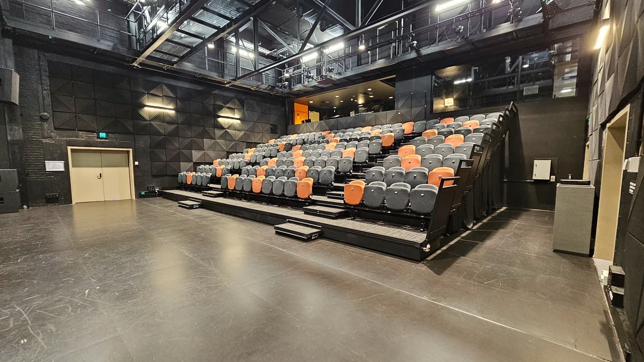 On the right, tiered seating features black and orange chairs separated by an aisle. To the left, a drinking fountain is present. An elevated area is equipped with chairs. The ceiling adopts an industrial look with exposed beams and fluorescent lights. The area has dimmed lighting, black wall tiles, complemented by grey tiles. A door on the left is marked with an Emergency Exit sign on top.