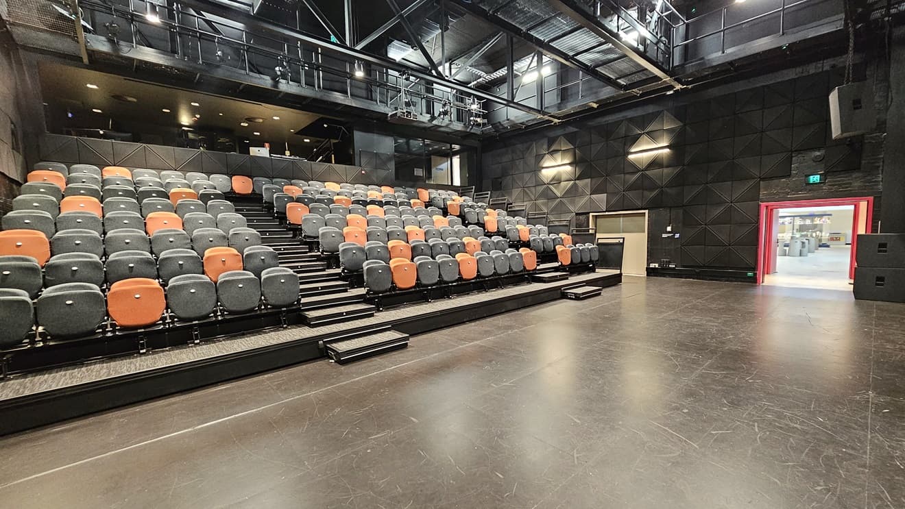 In the middle, tiered seating features black and orange chairs separated by an aisle. An elevated area is equipped with chairs. The ceiling adopts an industrial look with exposed beams and fluorescent lights. The area has dimmed lighting, and black wall tiles complemented by grey tiles. A bright pink door on the right has an Emergency Exit sign on top, and another white door to its left. The wall is soundproofed with black tiles.