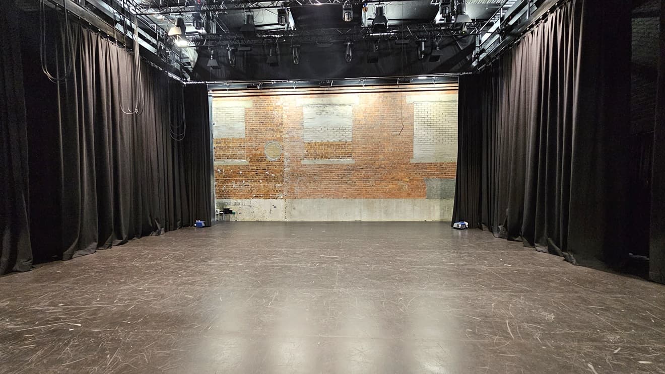 In the middle, there's a stage with a red brick wall backdrop. The stage area contains substantial theatre production equipment, featuring boom lighting and large black drop curtains against the left and right brick walls. The floor is composed of grey tiles.