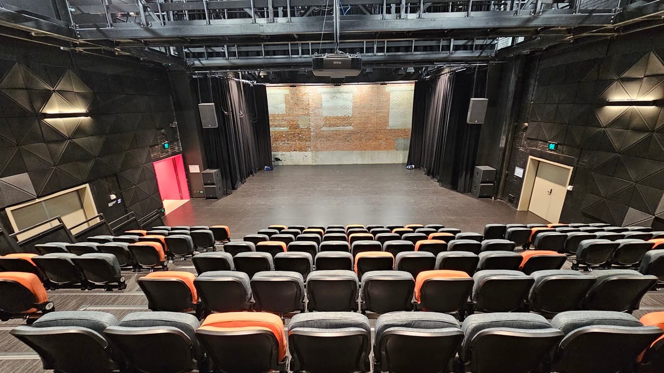 The tiered seating consists of orange and black chairs, with an aisle on the left dividing them. In front is the stage, featuring black drop curtains. A pink door is on the left, and a white door is on the right. The ceiling is adorned with industrial metal beams. Rubber tiles are affixed to the walls on both sides.