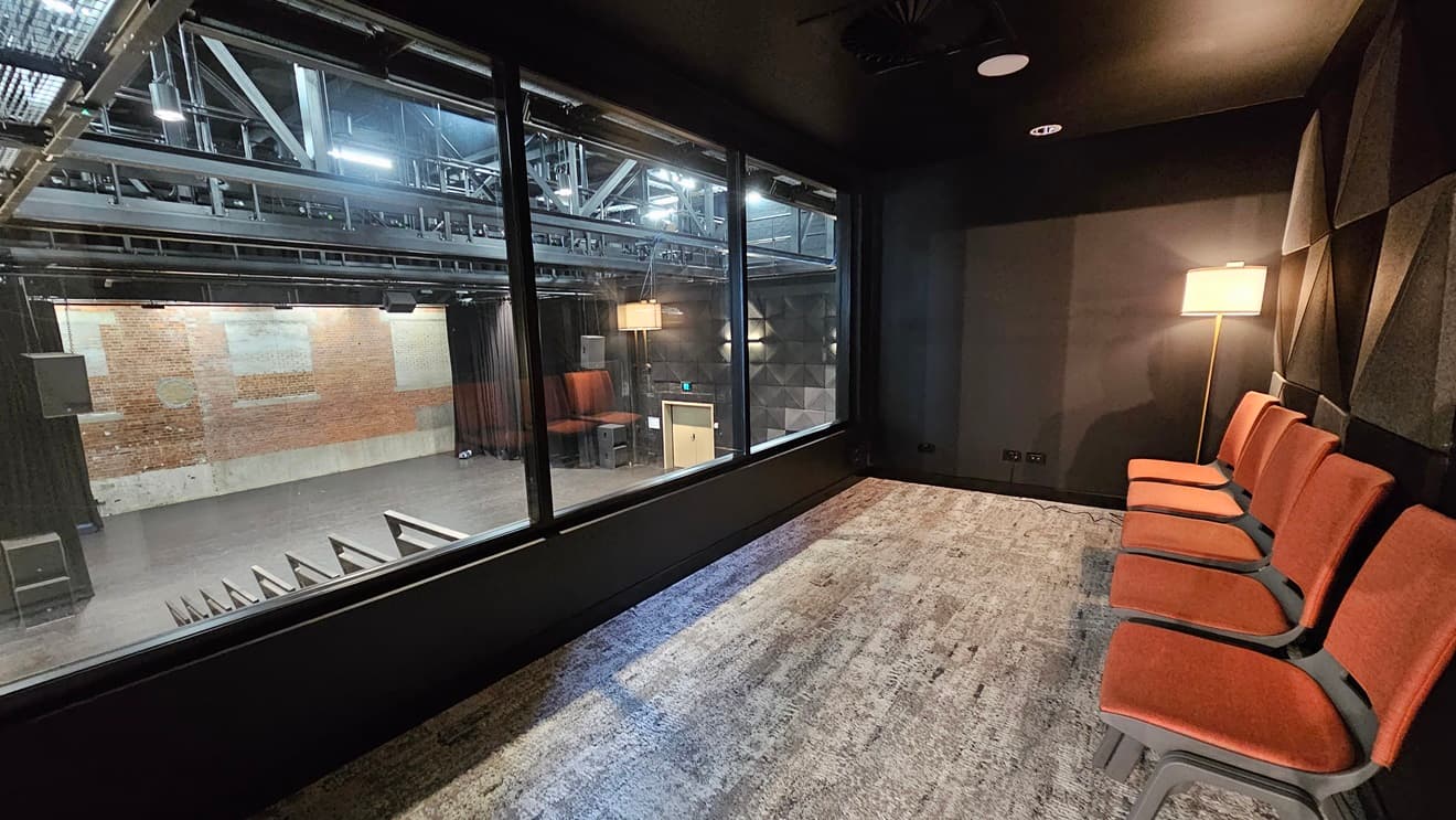 This room has orange chairs arranged on one side, enclosed by glass, overlooking the stage. The wall is surrounded by black rubber tiles, and the wall itself is black. The room is partially dim. In front is the stage with black drop curtains, and the ceiling features industrial metal beams. Rubber tiles are affixed to both sides of the walls.
