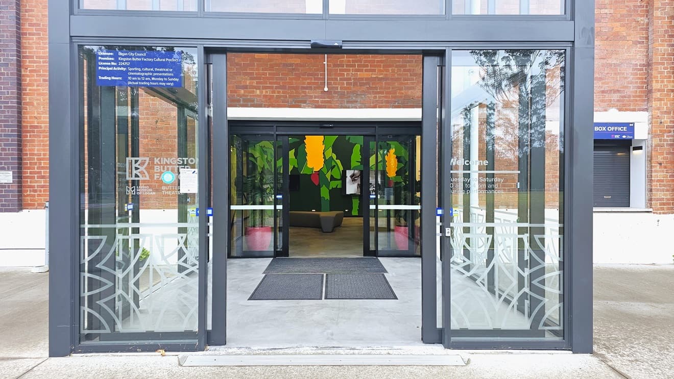 In the middle is a double glass door, furnished with non-slip mats and step free ramp. The front door displays the facility's name and store hours. Inside there is a flat-screen TV mounted on a colorful wall and a seating bench is positioned in the middle. Both sides show the building facade made of red brick walls. A window is visible on the left wall.