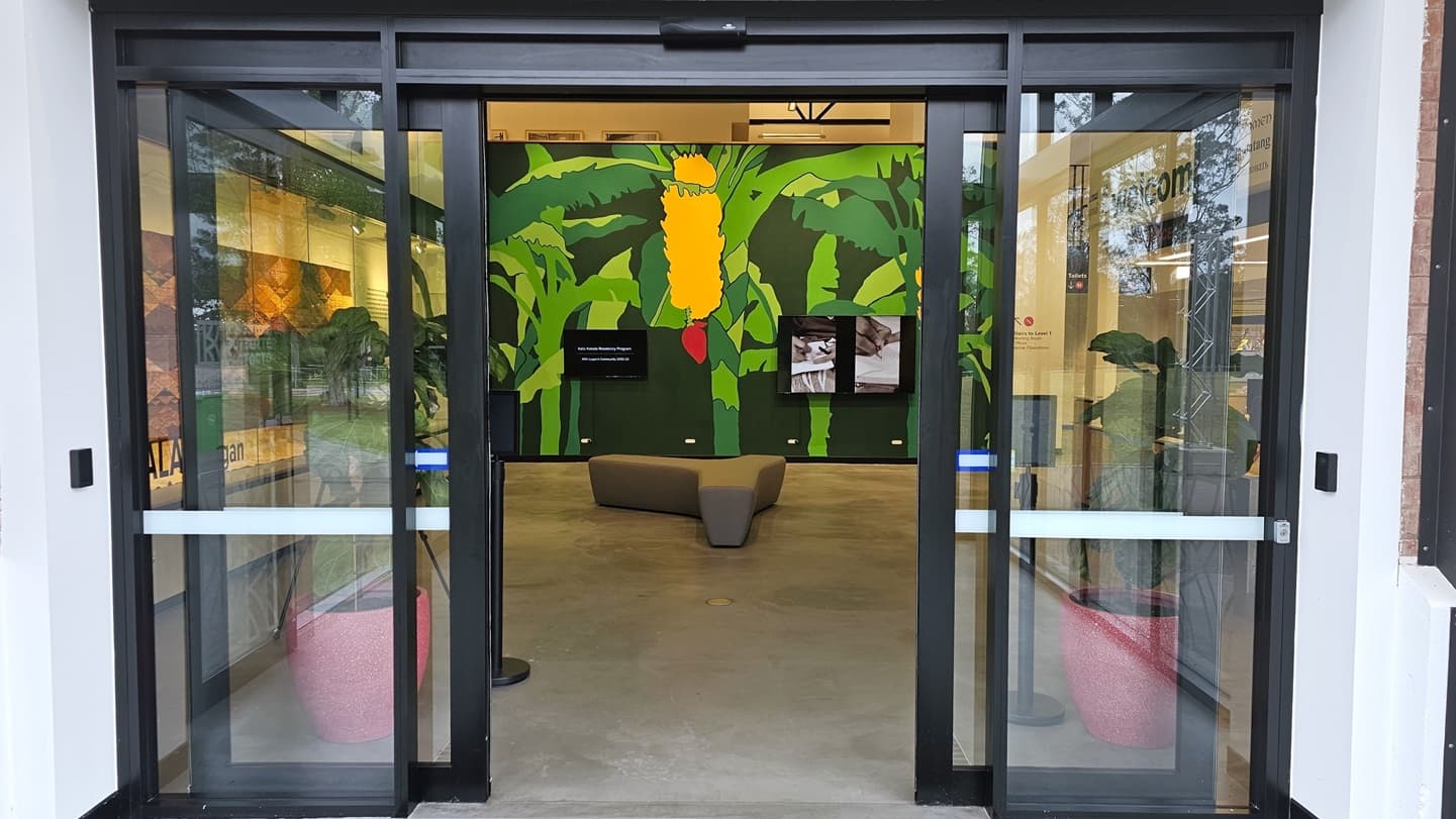 The entrance is an automatic glass sliding door with a white sticker lining. Inside, a Y-shaped cushioned chair is in the middle. A green wall featuring banana trees with fruit is present. Two flat screens are mounted. To the right is the reception. A pot of ornamental plants is positioned near the door entrance.