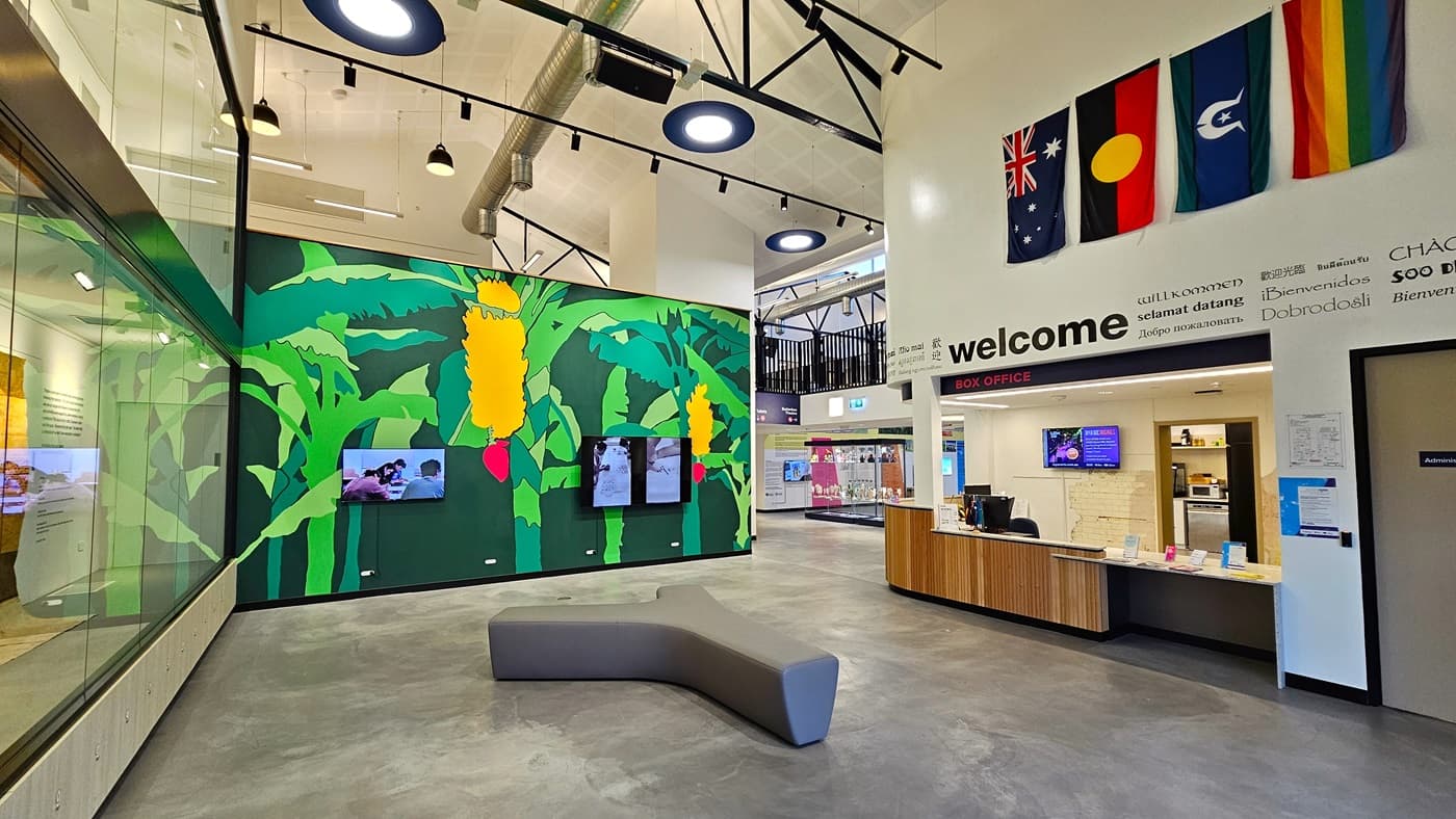 In the middle is a Y-shaped cushioned chair. A green wall featuring banana trees with fruit is present. To the right is the reception, adorned with a wooden desk displaying various posters and flags. Flat-screen TVs are also mounted on the middle wall and at the reception. Banners are affixed to the right wall. To the left is a floor-to-ceiling glass wall. The ceiling is adorned with circular light fixtures. On the far right, a section of the mezzanine is visible.