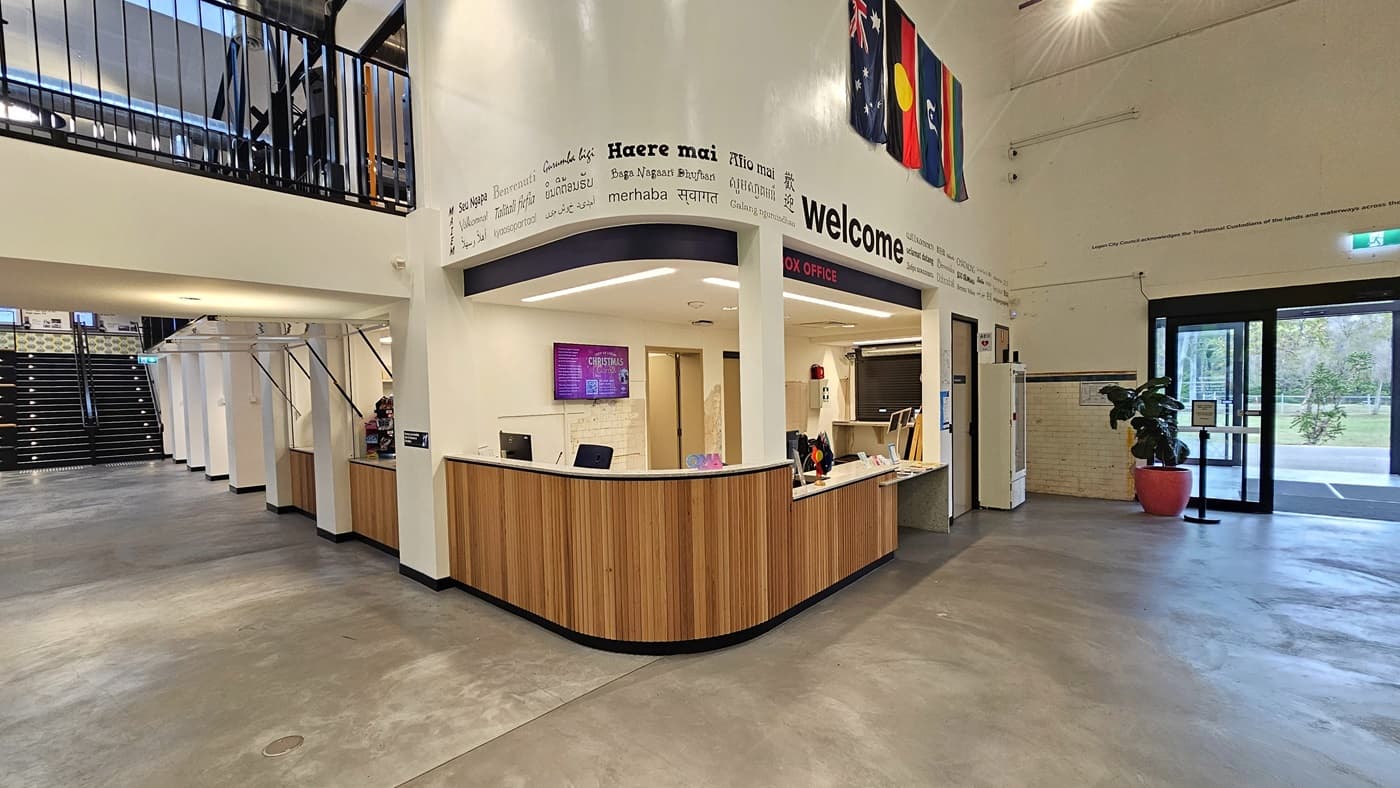 On the right is the reception, featuring a wooden desk with various flags. Flat-screen TVs are mounted on the middle wall of the reception. Banners and posters are affixed to the right wall. To the left is a staircase leading to the mezzanine. A section of the mezzanine is also visible. To the right is the door entrance, with a pot of ornamental plants positioned near it.