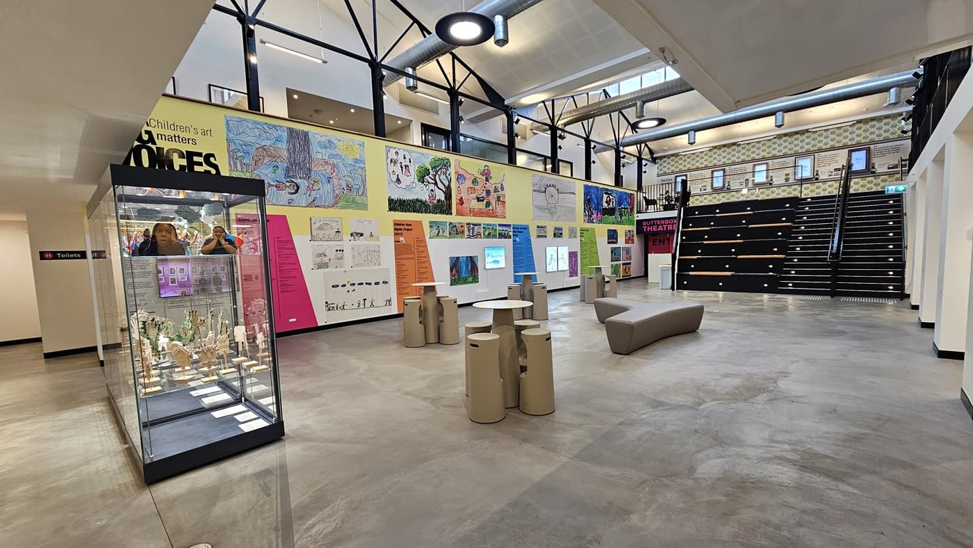 In the centre is the reception, alongside a small café. The desks in both the reception and café are designed with a wooden timber motif. Flat-screen TVs are mounted on the walls. To the left is an area leading to an open space with seating. The mezzanine level is also visible. The area is well-lit with light fixtures and fluorescent lights.