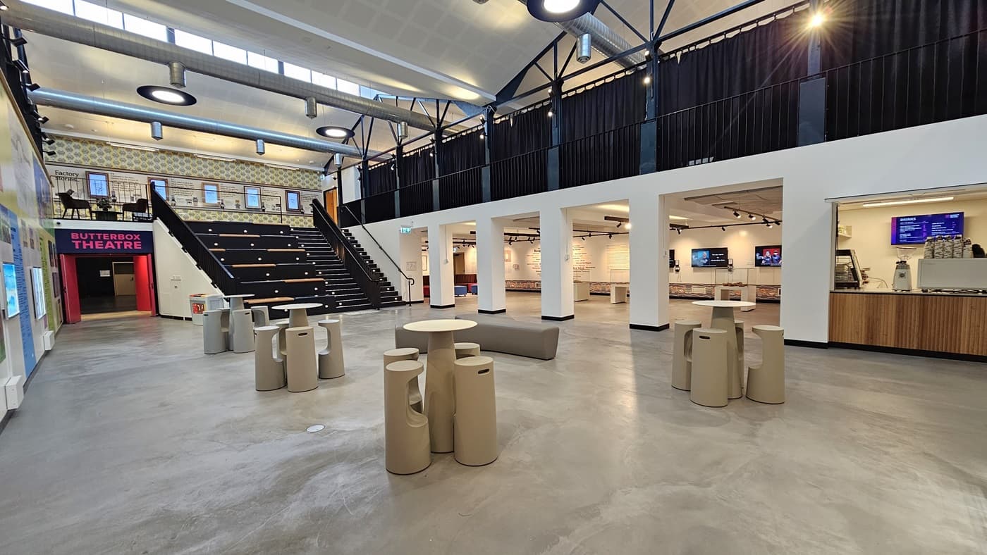 On the left, the wall displays various posters, paintings, and art materials. Bright pink entrance doors are situated on the left. In the middle, there are Y-shaped cream-cushioned chairs along with uniquely designed stools and tables. A stair leading to the mezzanine and tiered seating is also positioned in the middle. The ceiling features an open space with circular lights and large air-conditioning vents. The cafe is on the right, and further ahead is a spacious seating area.