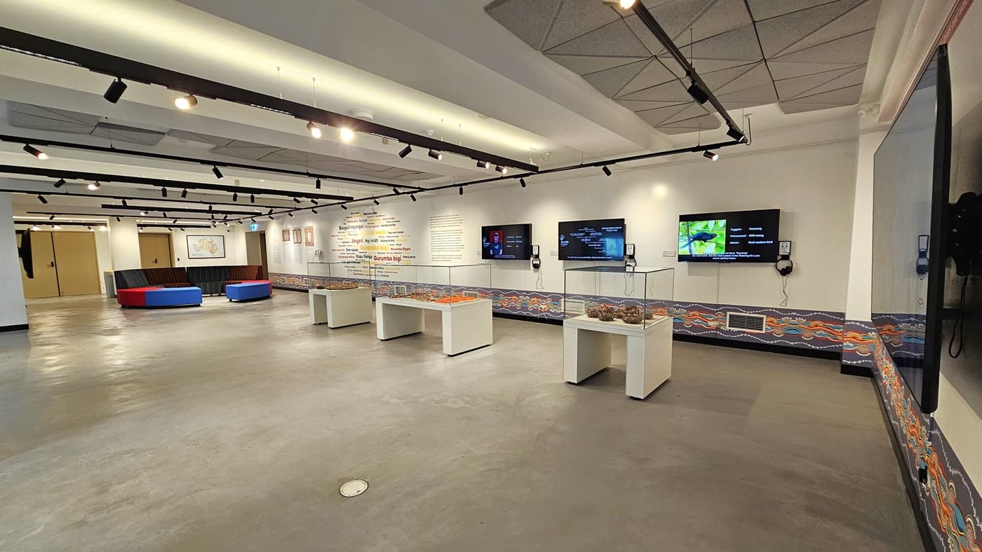 On the right is a spacious area with cushioned seating in the corner. There are three tables with enclosed glass displays. The right wall features meticulously painted wall art. Large flat-screen TVs are mounted on the wall. The ceiling is white with light fixtures and ceiling art decoration. The floor area is made of polished concrete.