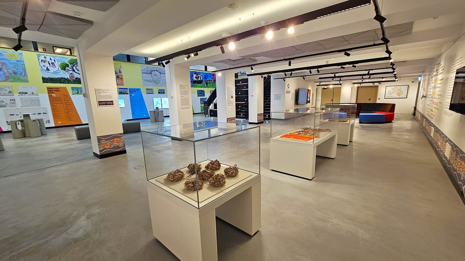 On the right is a spacious area with cushioned seating in the corner. Three tables with enclosed glass displays are present. The right wall showcases meticulously painted wall art, with large flat-screen TVs mounted on it. The ceiling is white, adorned with light fixtures and ceiling art decoration. The floor area is made of polished concrete. The right wall is adorned with art posters and paintings, and there are uniquely designed stools and tables for seating.
