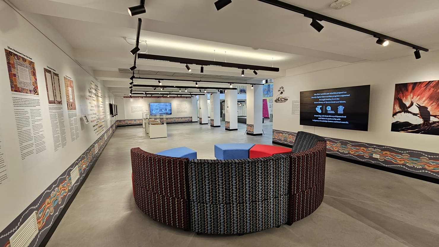 The entire wall showcases meticulously painted wall art and art posters. Large flat-screen TVs are mounted on both the right and middle sections of the wall. The ceiling is white, adorned with light fixtures and ceiling art decoration. The floor area is made of polished concrete. In the middle, there is a cushioned seating area arranged in a semi-circle layout.
