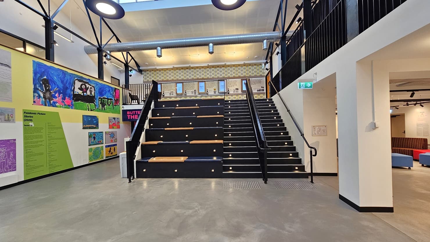 In the centre is a staircase fitted with truncated columns, leading to the mezzanine. To the left, there's a tiered seating area. The left wall is adorned with art images and paintings, with entry to the Butter Theatre on the left. On the right is a seating area with a couch. At the top of the stairs, the mezzanine wall adorned with framed posters is visible.
