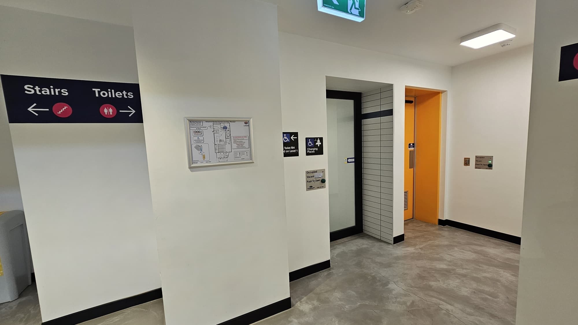 To the left, a wall features directional signage. On the right, is an accessible bathroom and changing place, marked with gender and accessibility signs. The right bathroom has a yellow swing door, while the middle one has a manual white sliding door. A framed poster on the left displays the facility map. The wall is white with tile accents.