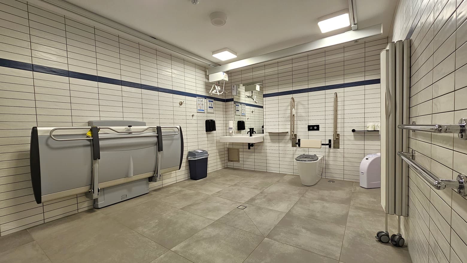 Within the accessible bathroom, an adult-sized change table is affixed to the left wall. A collapsible wall divider stands on the right. In the middle, an ambulant toilet bowl, featuring handrails, is positioned. On the wall, a washbasin, liquid soap dispenser, and tissue dispenser are installed. Tissue rolls are organized on the right, with rubbish bins on both sides. The entire wall is tiled in white, and an additional hand grab is mounted on the right wall.