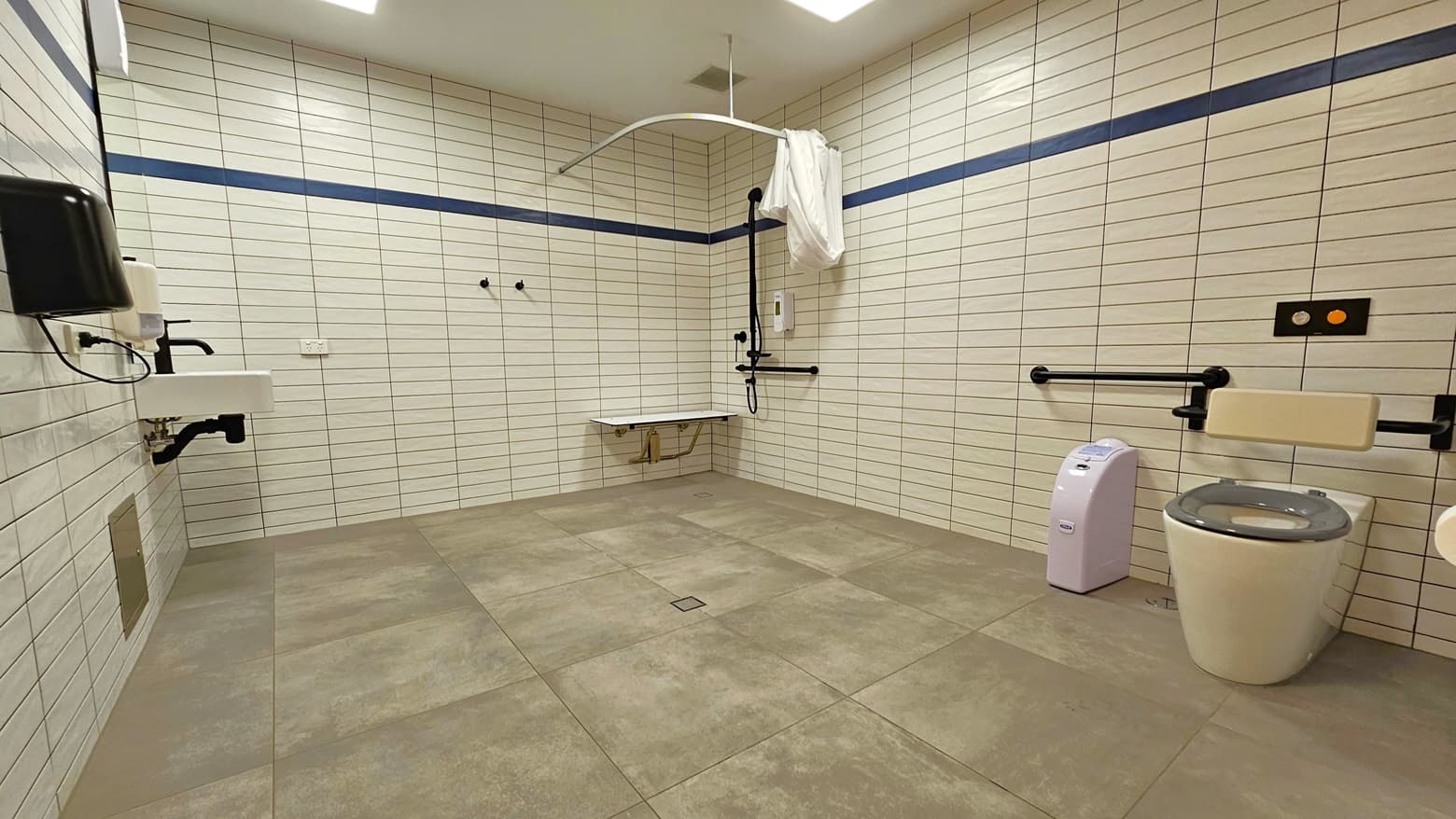 In this spacious bathroom, a shower is situated in the right corner, accompanied by handrails, a bench chair, and a hoist. On the right, a toilet bowl with handrails and a rubbish bin is present. To the left, a washbasin is affixed to the wall, along with a liquid soap dispenser and a hand dryer. The entire area is surrounded by white tiles and white ceilings, while the floor is matte grey.