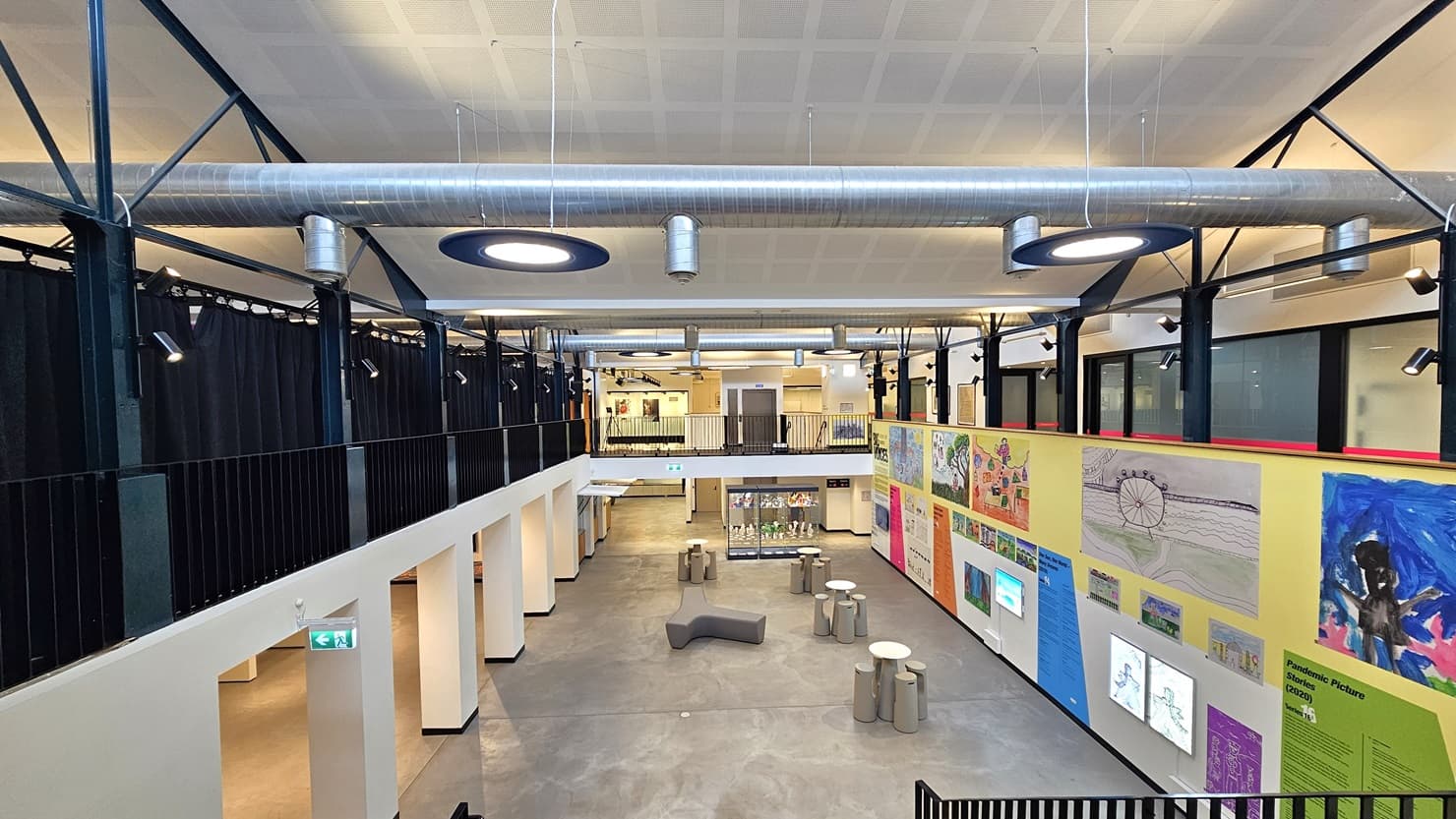 The mezzanine provides a view of the spacious lobby below, featuring a wall adorned with art paintings. Tables and chairs are arranged in the middle, accompanied by a glass display. Navy blue curtains are on the left with low iron railings. To the right, there is another lobby leading to function rooms. The ceiling is white with exposed air-conditioning vents, and stylish circular lights hang from above.