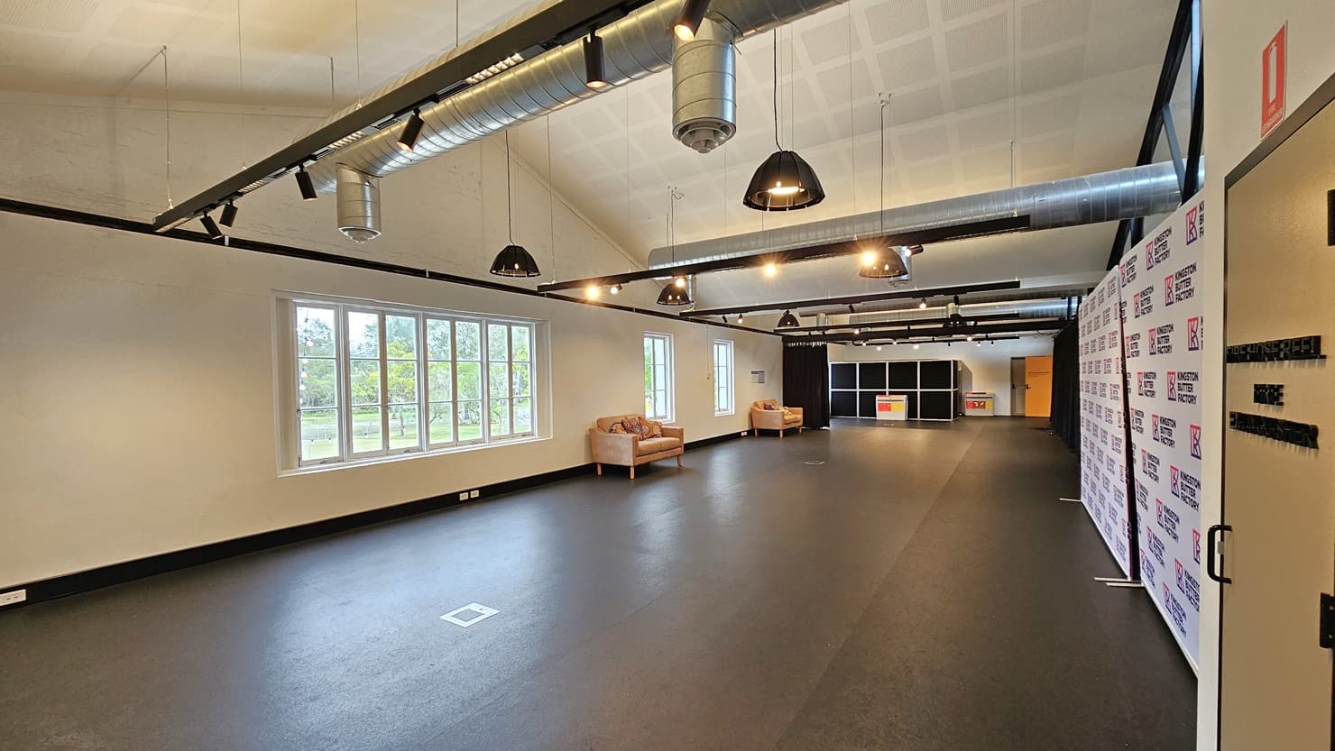 A detailed view of the expansive room reveals cushioned chairs on the left, near large glass windows. On the right, an area is enclosed with navy blue curtains and retractable barriers, featuring a black display rack in the distance. The ceiling is white with exposed air conditioning vents, and light fixtures hang from the white wall. The floor is dark grey.