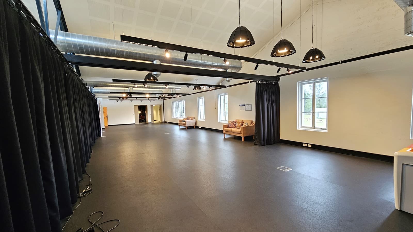 Examining the expansive room, cushioned chairs line the right side near large glass windows. On the left, navy blue curtains and retractable barriers are present. Two doors are situated at the far end. The ceiling is white with exposed air conditioning vents, and light fixtures hang from the white wall. The floor is dark grey, with electrical cords visible on the left floor.