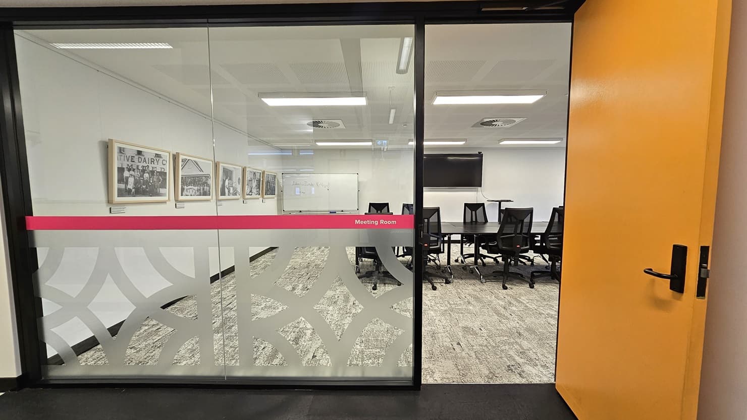 To the left is a glass wall with white and pink print stickers indicating "meeting room." To the right is a yellow wooden door. Inside, a long black conference table is surrounded by black swivel chairs. The left wall features framed posters, and there's a large whiteboard and a flat-screen TV in the middle. The floor has grey carpet, and the ceiling is entirely white with fluorescent lights and centralized air conditioning.