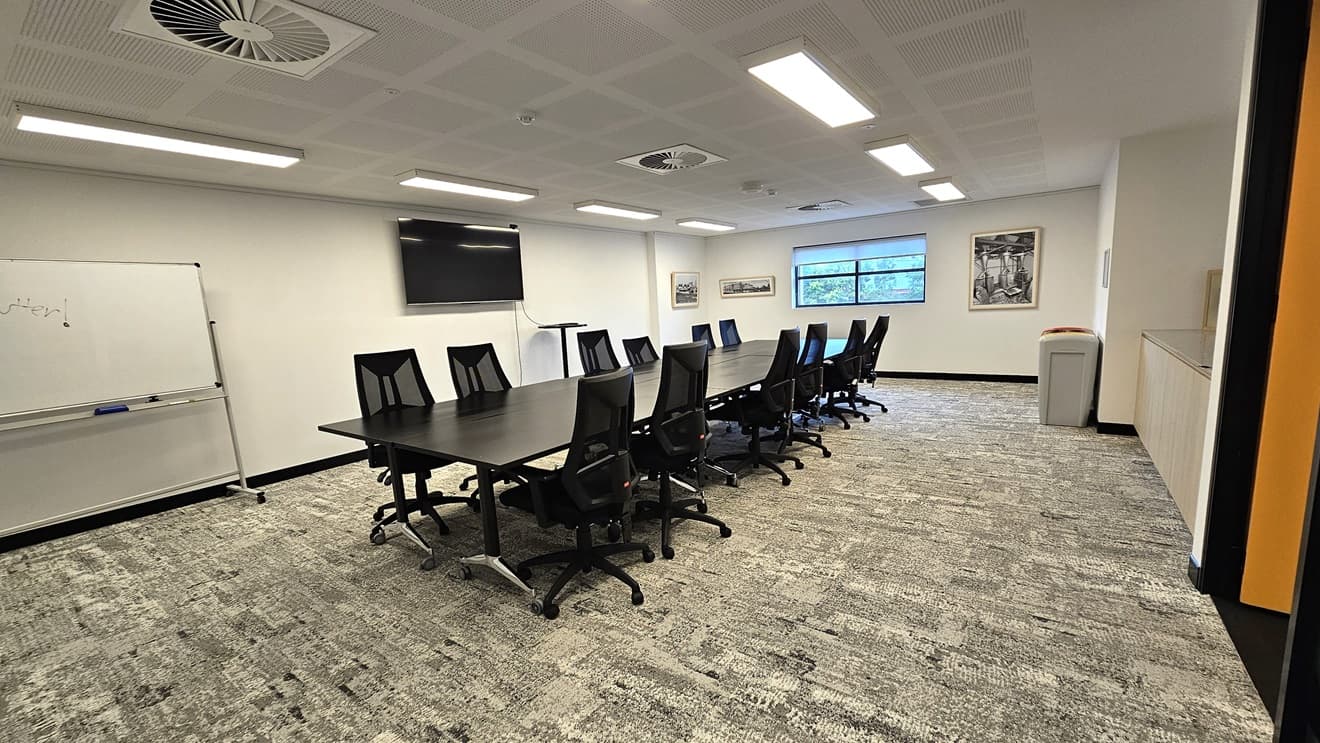 A detailed look at the meeting room reveals a long black conference table in the middle, surrounded by black swivel chairs. The central wall has a large glass window and framed posters. On the left wall, there's a large whiteboard and a flat-screen TV. The floor is covered with grey tiles, and the ceiling is entirely white, featuring fluorescent lights and centralized air conditioning.