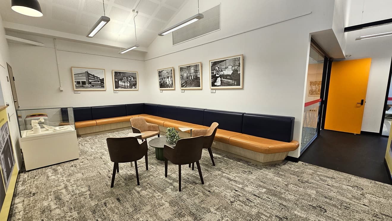 The seating area comprises an L-shaped black and brown couch with a central table surrounded by seating, adorned with a potted plant. The wall is decorated with glass-framed posters, and on the left is a table with glass panels. The floor is covered with grey carpet, and the ceiling is entirely white, featuring fluorescent lights and centralized air conditioning. On the right, there is a glass wall and a yellow door.