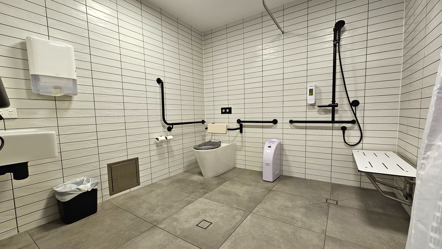 The spacious bathroom includes a corner toilet with handrails and a mounted black shower. A tissue dispenser is located beside the toilet. On the right, a wall-mounted bench chair and a white curtain are positioned. To the left, a washbasin and another tissue dispenser are available, with a rubbish bin underneath. The walls are covered in white tiles, and the ceiling is white. The floor is tiled in grey.