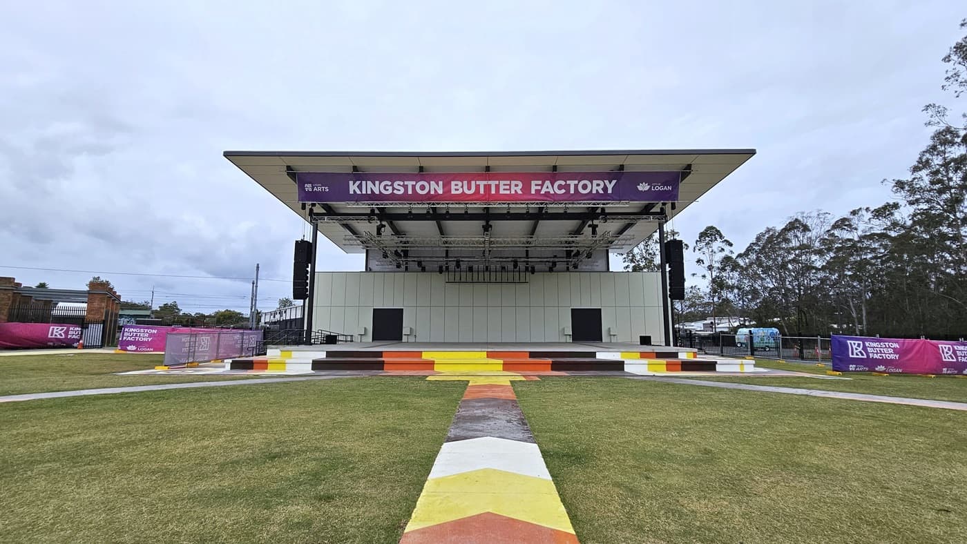 A concrete pathway runs across the grassy area toward the stage. The stage has colorful steps and white wall tiles. A gradient pink and blue banner displaying the facility's name hangs near the ceiling. On the left, the Butter Factory building is present with a red brick facade and gradient banners. Picnic tables are positioned in the grassy area.