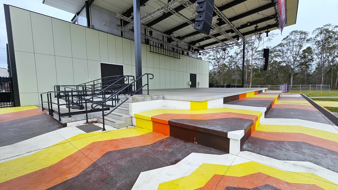 The stage steps are adorned with colourful lines in yellow, orange, brown, and white. To the left, there are additional entries, including stairs and a ramp with handrails and truncated domes. The stage exhibits white walls and an industrial ceiling with exposed beams. Large audio speakers are suspended on both sides of the stage. The surroundings include trees and grass.