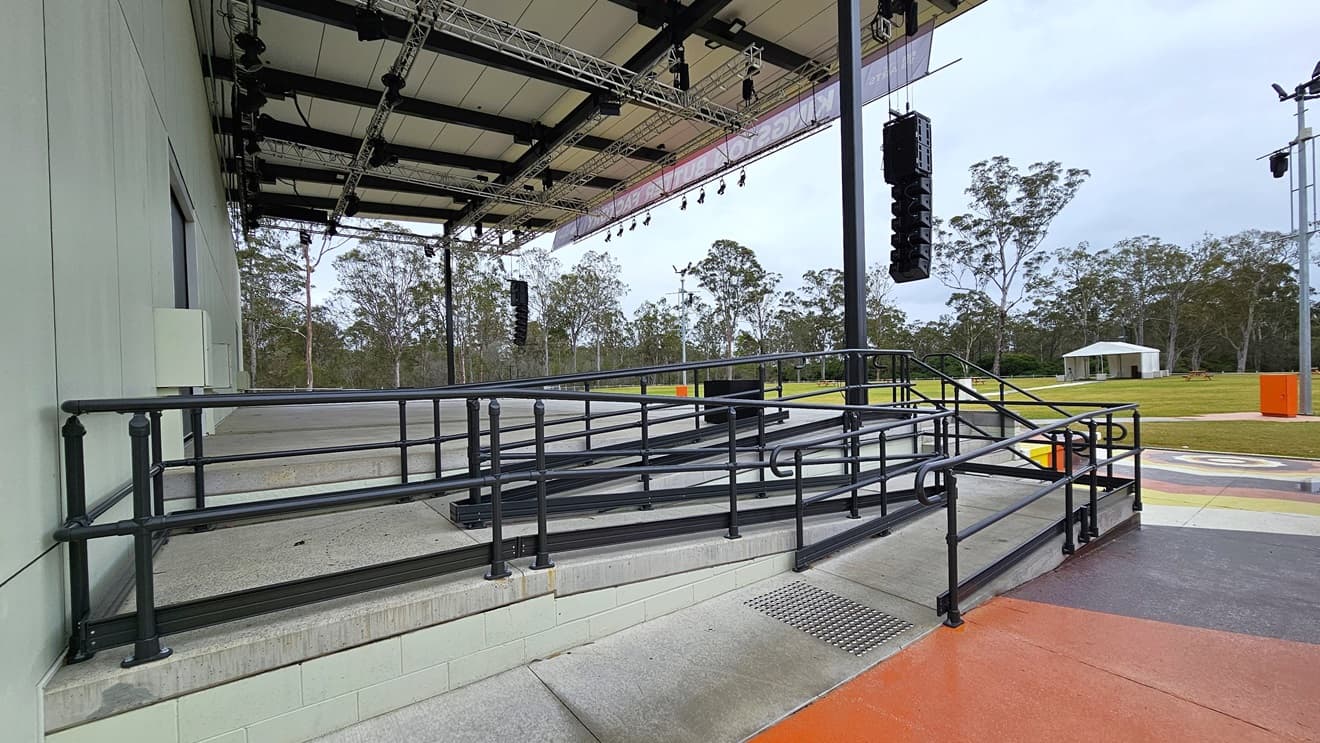 A wheelchair-accessible entry is situated on the left of the stage, featuring a ramp with handrails and truncated domes on the floor. The stage displays white walls and an industrial ceiling with exposed beams. Large audio speakers are suspended on both sides of the stage. Colourfully painted steps adorn the front of the stage, and a lamp post stands in the grassy area.