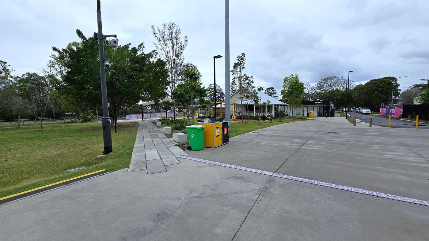A concrete path extends to the grassy area, bordered by landscaping and trees on the right. Additional trees line the left side, while tall light posts and building structures stand in the middle. On the right, a concrete road leads to the car park, bordered by yellow truncated domes.