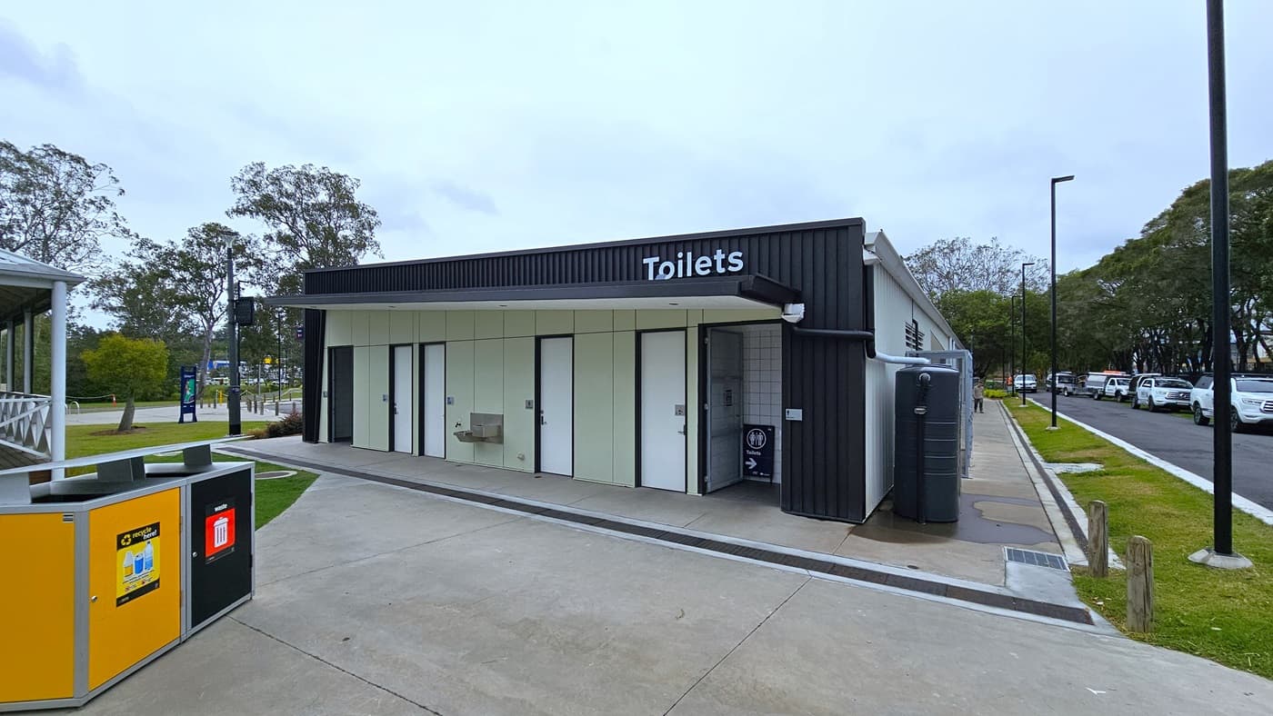 A toilet block, featuring 6 white doors marked for gender access, is situated near the road. The building has corrugated design black walls. On the left, yellow and black rubbish bins are marked for separating recyclables. A section of a building structure is on the left, while to the right, a road accommodates a few parked cars.