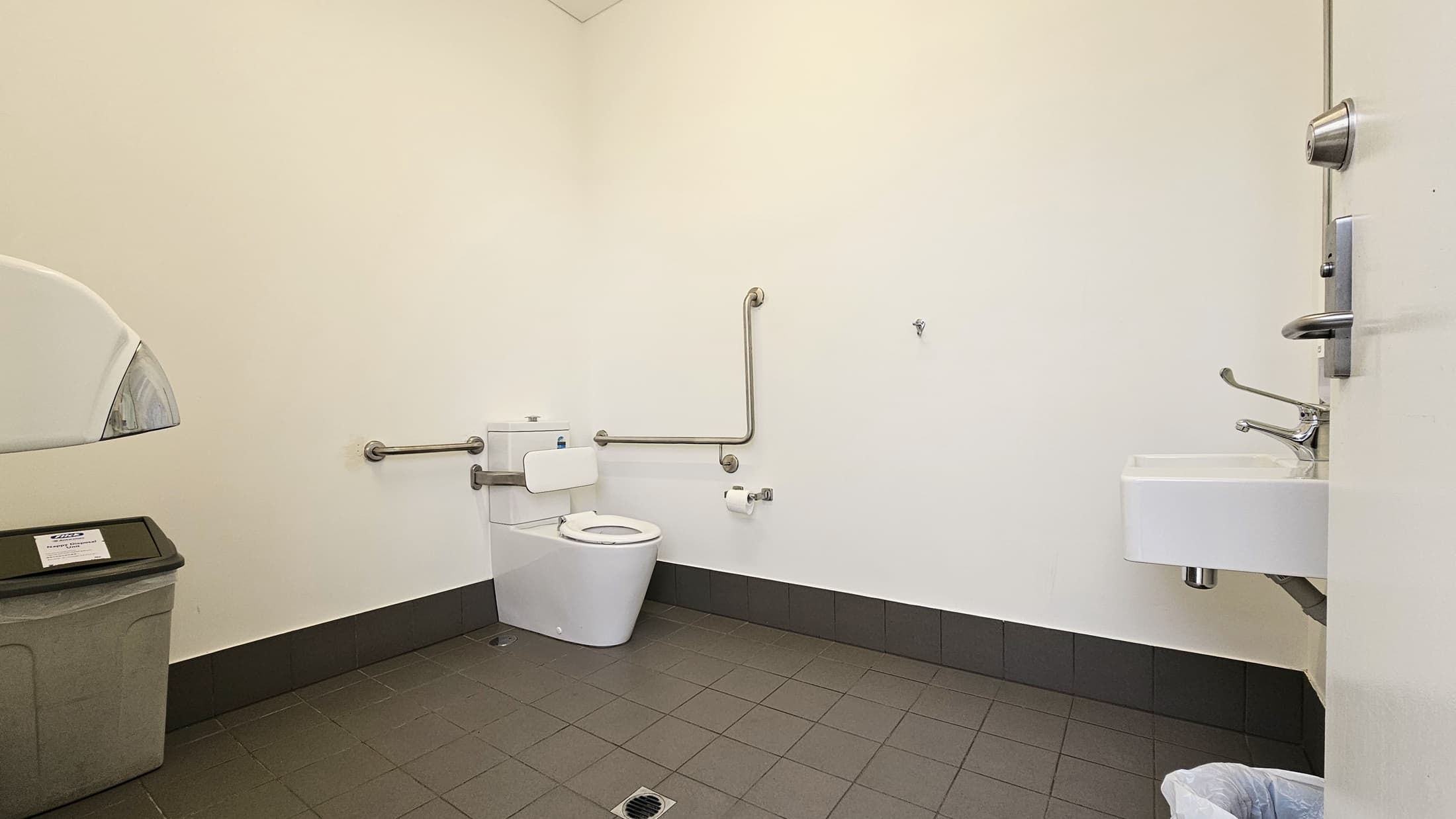 Inside the toilet, a toilet bowl equipped with handrails is positioned in the corner along with a tissue dispenser. To the right, a washbasin is mounted, and a rubbish bin is placed underneath. On the left, a tissue dispenser is mounted to the wall with a rubbish bin underneath. The wall is white, and the floor has grey tiles.