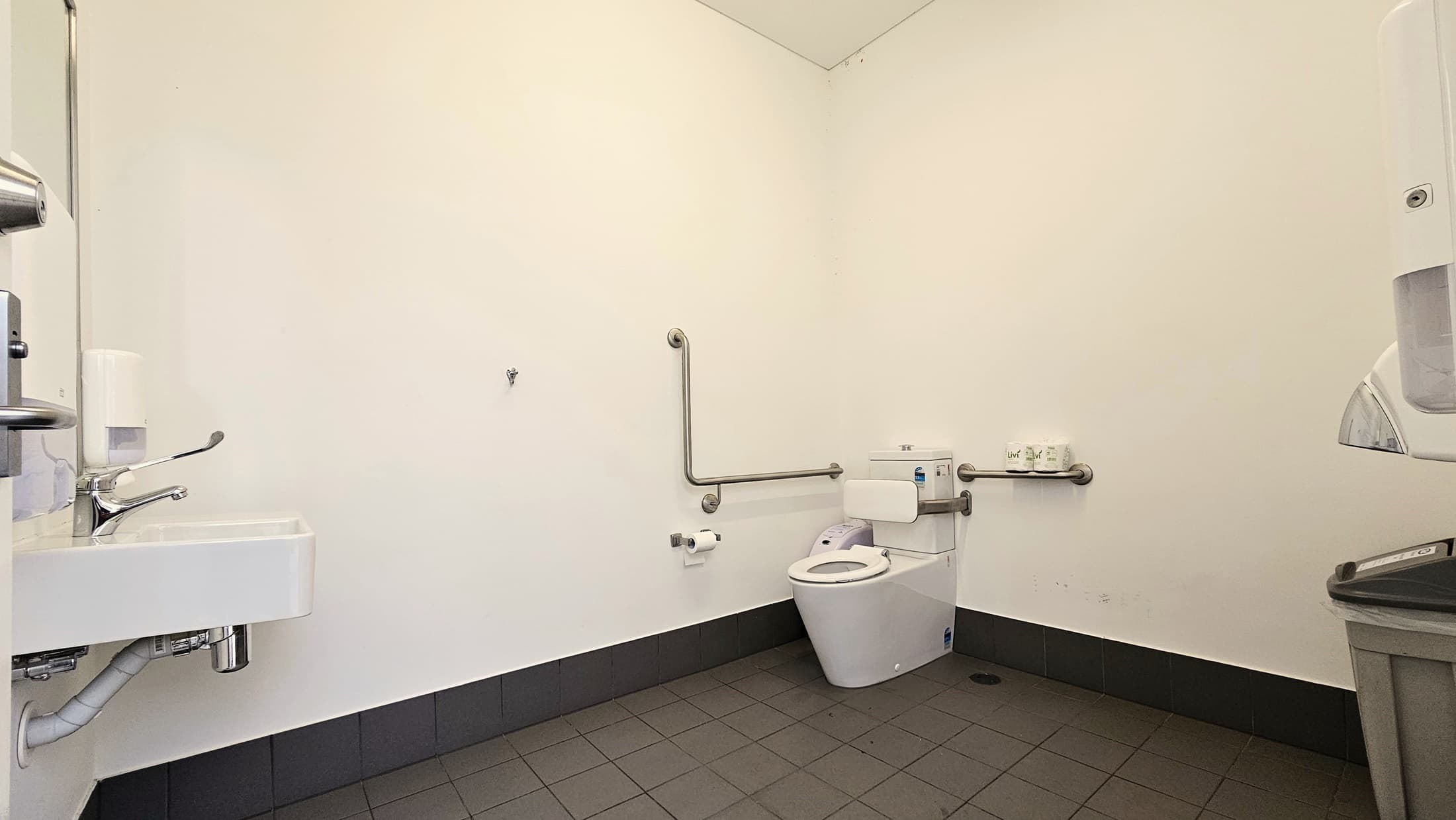 Inside the toilet, a toilet bowl equipped with handrails is positioned in the corner, accompanied by a tissue dispenser. On the left, a washbasin with a liquid soap dispenser is mounted on the wall, and a rubbish bin is placed underneath. On the right, a tissue dispenser is mounted to the wall with a rubbish bin underneath. The wall is white, and the floor has grey tiles.