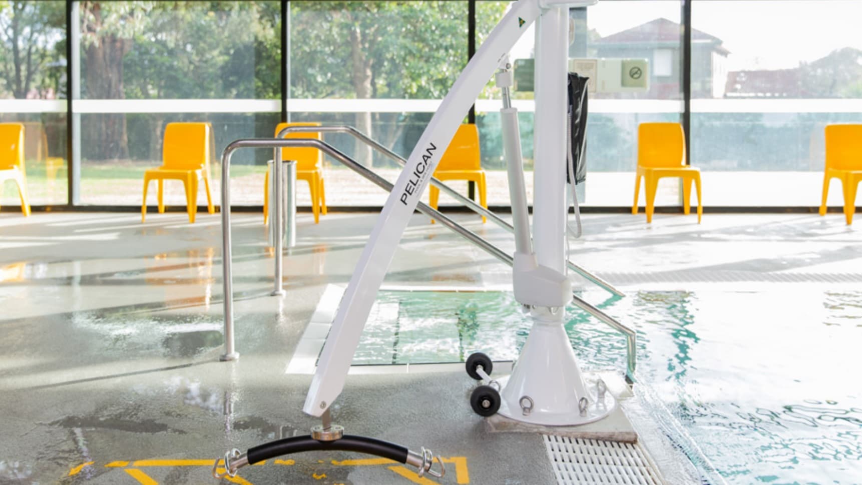 A poolside area with a white mechanical lift labelled "PELICAN" positioned near the water's edge. The floor is wet and reflective. In the background, there are several bright orange chairs lined up against large glass windows, which offer a view of greenery outside. The pool has metal handrails leading into the water.