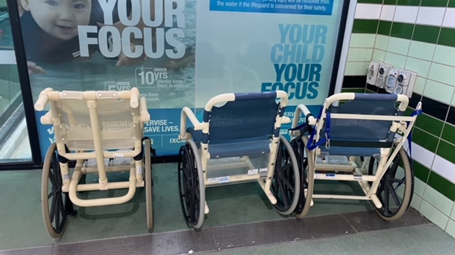 Three wheelchairs are lined up side by side against a wall. The wheelchairs have white frames and blue seats. Behind them, there is a large advertisement poster featuring a child's face and text. The wall is tiled with a pattern of green, white, and black tiles. On the right, there is a small control panel or device attached to the wall. The floor is grey and smooth.