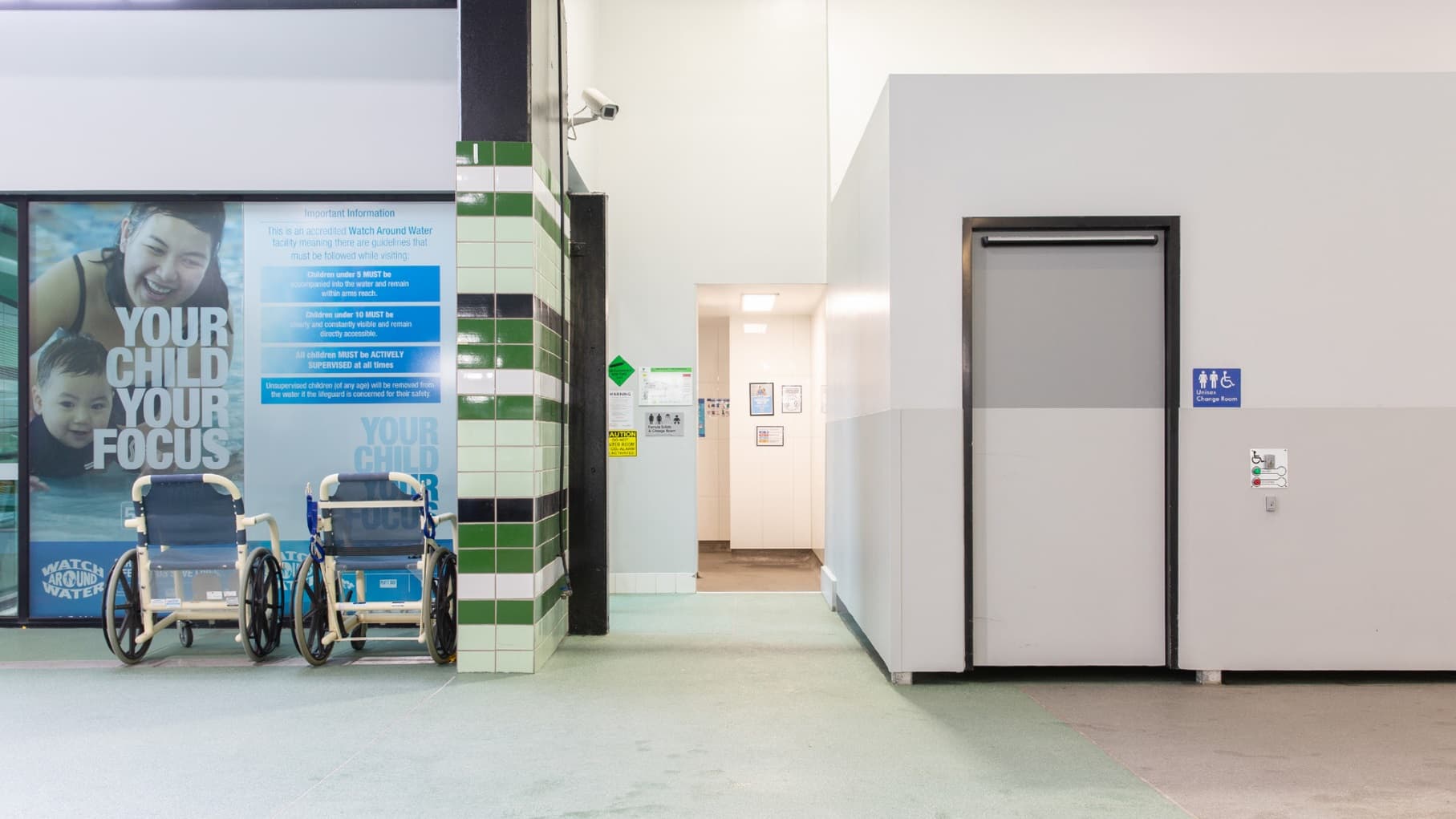 On the left, there are two wheelchairs in front of a large poster featuring a child and an adult with the text "YOUR CHILD YOUR FOCUS." The wall has green and white tiles. In the centre, there's a corridor with a lighted interior and framed pictures on the wall. On the right, a grey door with a sign indicating accessible toilets. The floor is light-coloured, and the ceiling is white.