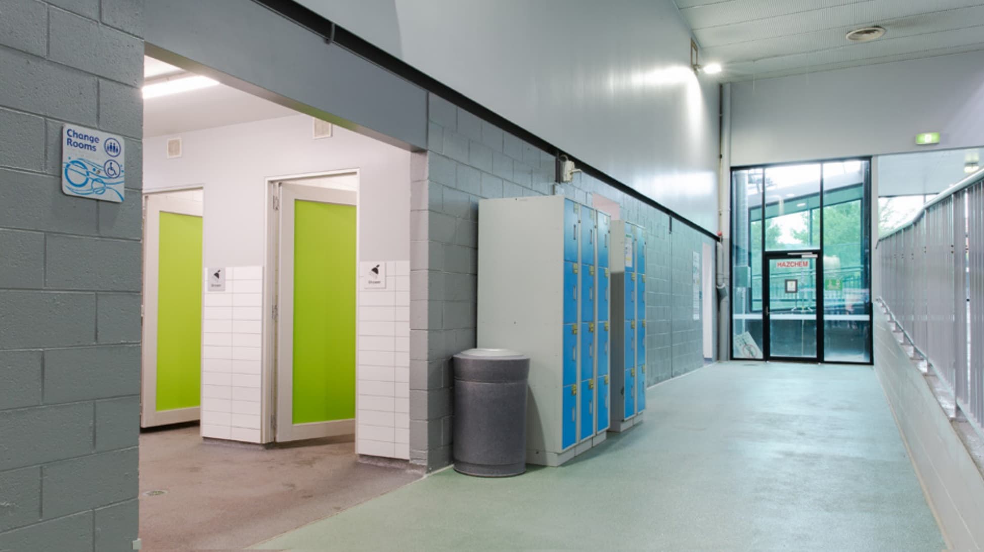 A corridor with light grey walls and a green floor. On the left, there are changing rooms with green doors and a sign indicating "Change Rooms". Adjacent to the changing rooms is a grey rubbish bin and a row of lockers with blue doors. On the right, a glass door leads outside, with a sign above it. The ceiling is white with fluorescent lighting.