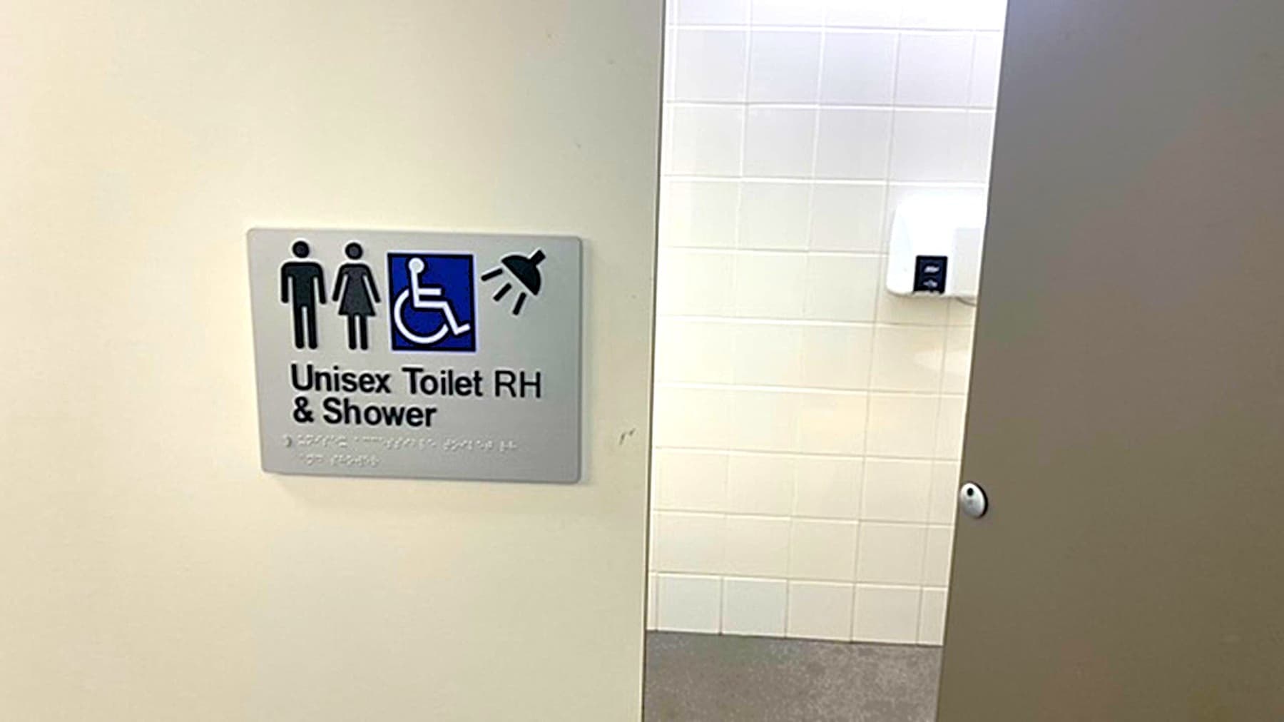 A sign on a wall indicates a unisex toilet with right-hand access and a shower, featuring symbols for male, female, wheelchair access, and a shower. The wall is light-coloured. To the right, a partially open door reveals a glimpse of the interior, showing a white wall and a mounted paper towel dispenser. The floor is grey.