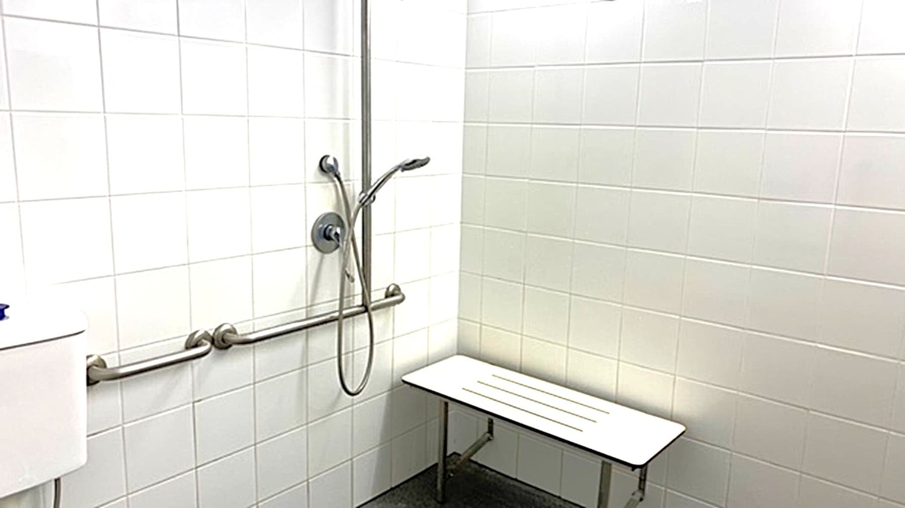 A bathroom with white tiled walls and a dark floor. On the left, there is a shower area with a handheld showerhead attached to a vertical rail, and a horizontal grab bar beneath it. A white toilet tank is partially visible. On the right, a light-coloured bench is fixed to the wall, supported by metal legs. The ceiling is not visible.