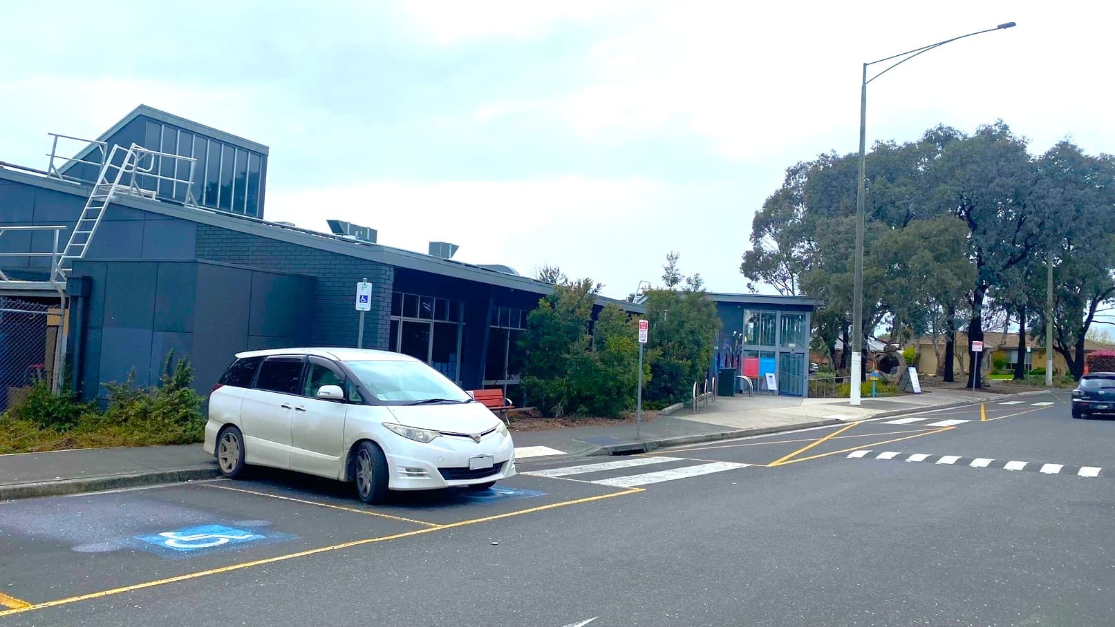 A white car is parked in one of the accessible parking spaces beside the sidewalk and directly opposite the main entry point of Knox Leisureworks. At the individual parking spaces is an accessible parking sign for persons with disabilities.