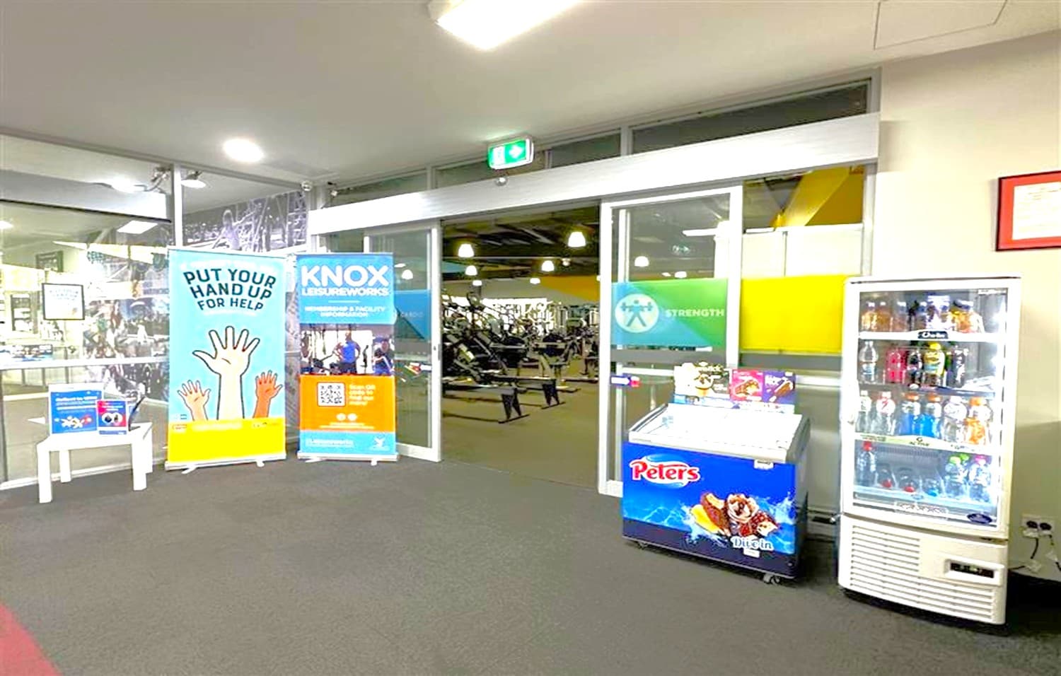 A gym entrance with glass doors in the centre, leading to a workout area with exercise equipment visible inside. On the left, there are two standing banners, one with a "Put Your Hand Up for Help" message and another for "Knox Leisureworks". A small white table holds additional pamphlets. On the right, a refrigerated display case with drinks and a freezer with "Peters" branding. The floor is carpeted, and the ceiling has recessed lighting.