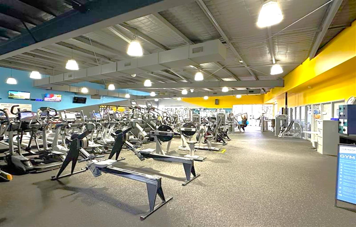 A spacious gym interior with a variety of exercise equipment. On the left, there are rows of treadmills and elliptical machines. In the centre, rowing machines are aligned. The right side features weight machines and a person using one. The walls are painted blue and yellow, with several mounted televisions. The ceiling is high with exposed beams and hanging lights. The floor is covered with dark, speckled rubber matting. A signboard is visible in the foreground on the right.