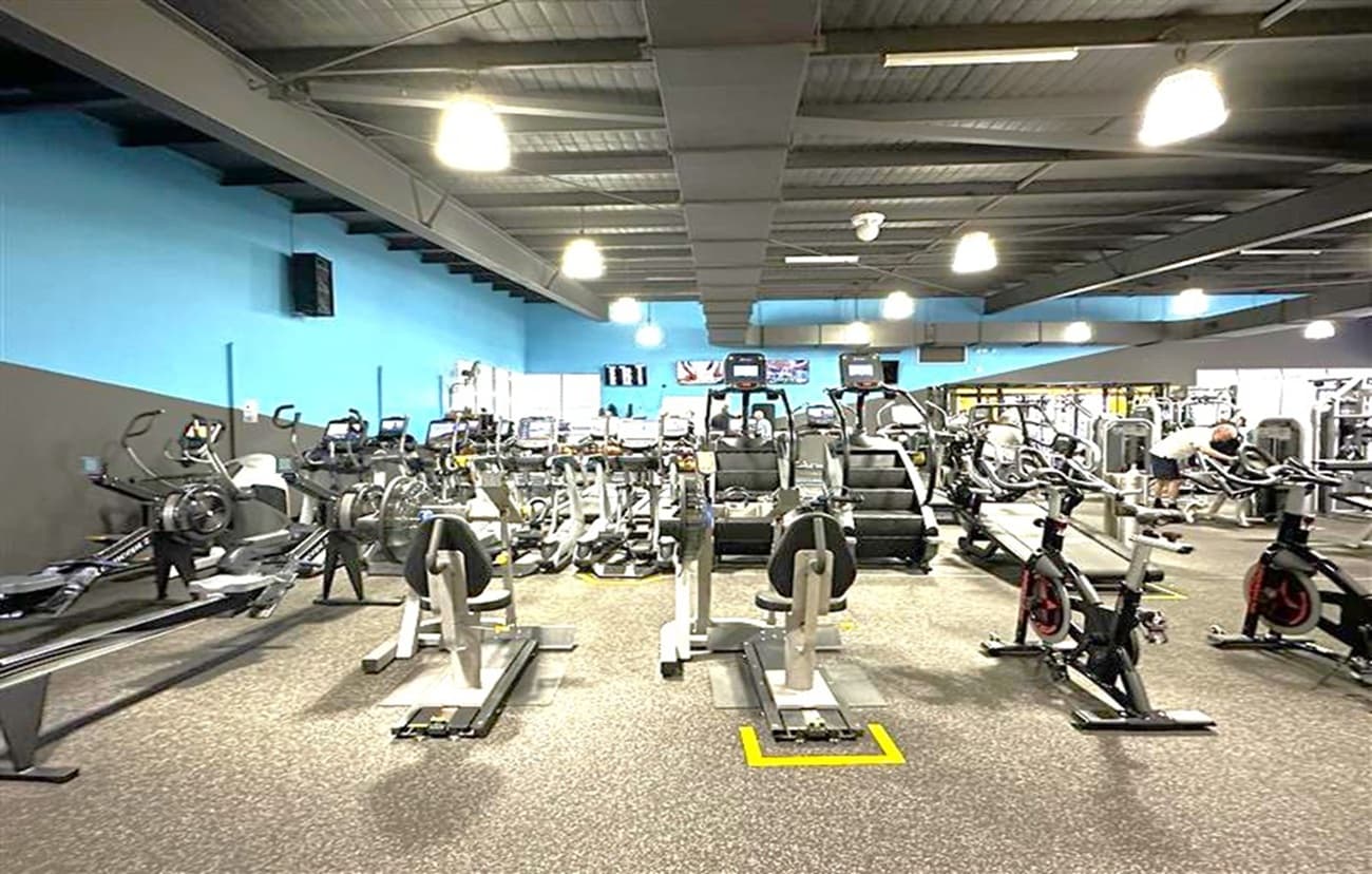 A gym interior with various exercise equipment. In the foreground, there are several rowing machines and stationary bikes. The middle section features treadmills and elliptical machines. The background shows more gym equipment and a person using a machine. The walls are painted blue, and the ceiling is industrial with exposed beams and hanging lights. The floor is covered with a speckled rubber mat.