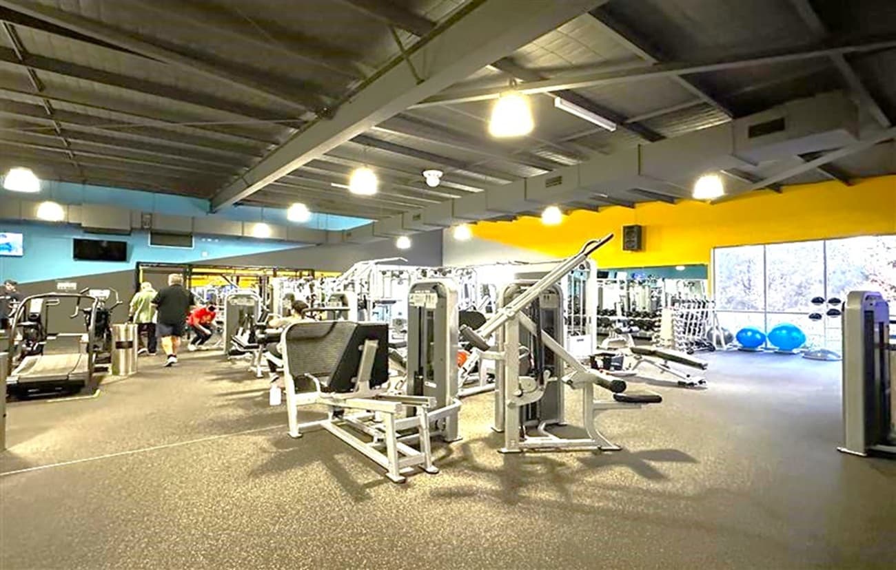 A spacious gym interior with various exercise machines and equipment. On the left, there are treadmills and people working out. In the centre, there are weight machines and benches. On the right, large windows allow natural light in, with blue exercise balls visible near the windows. The ceiling is high with exposed beams and hanging lights, and the floor is covered with dark carpeting. The walls are painted in a mix of blue and yellow.