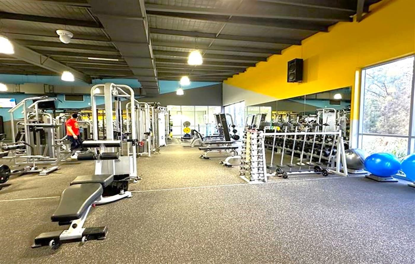 A gym interior with various exercise equipment. On the left, there is a bench press and a person in a red shirt near weight machines. In the centre, there are more weight machines and benches. On the right, a rack of dumbbells is positioned in front of a large mirror, with exercise balls on the floor nearby. The floor is covered with rubber mats, and the ceiling has exposed beams with hanging lights. Large windows on the right let in natural light.